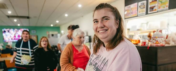 A smiling young woman in a pink sweatshirt during a community outing, with staff and other members behind her